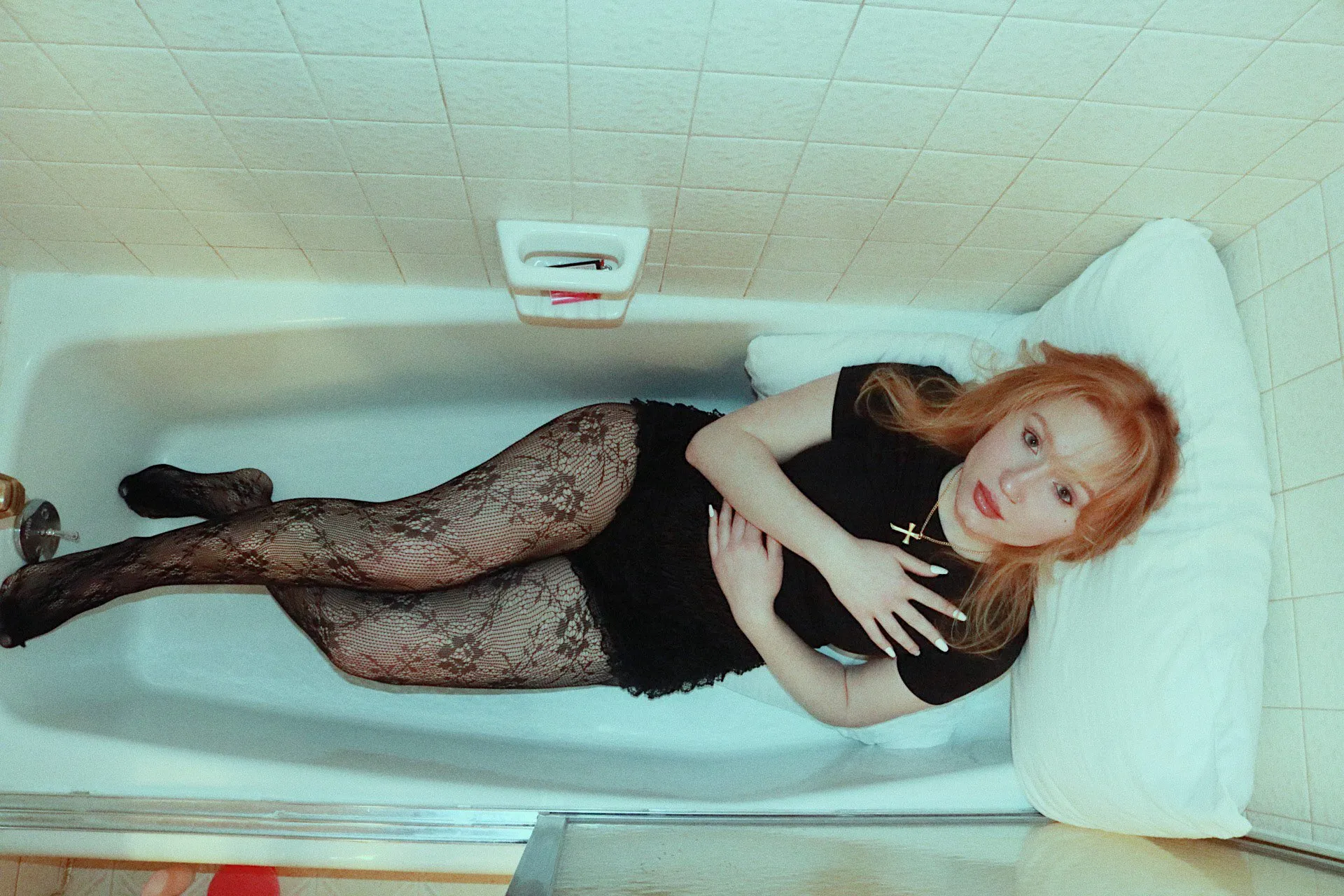 A woman in lace tights and a black top lies in an empty tiled bathtub