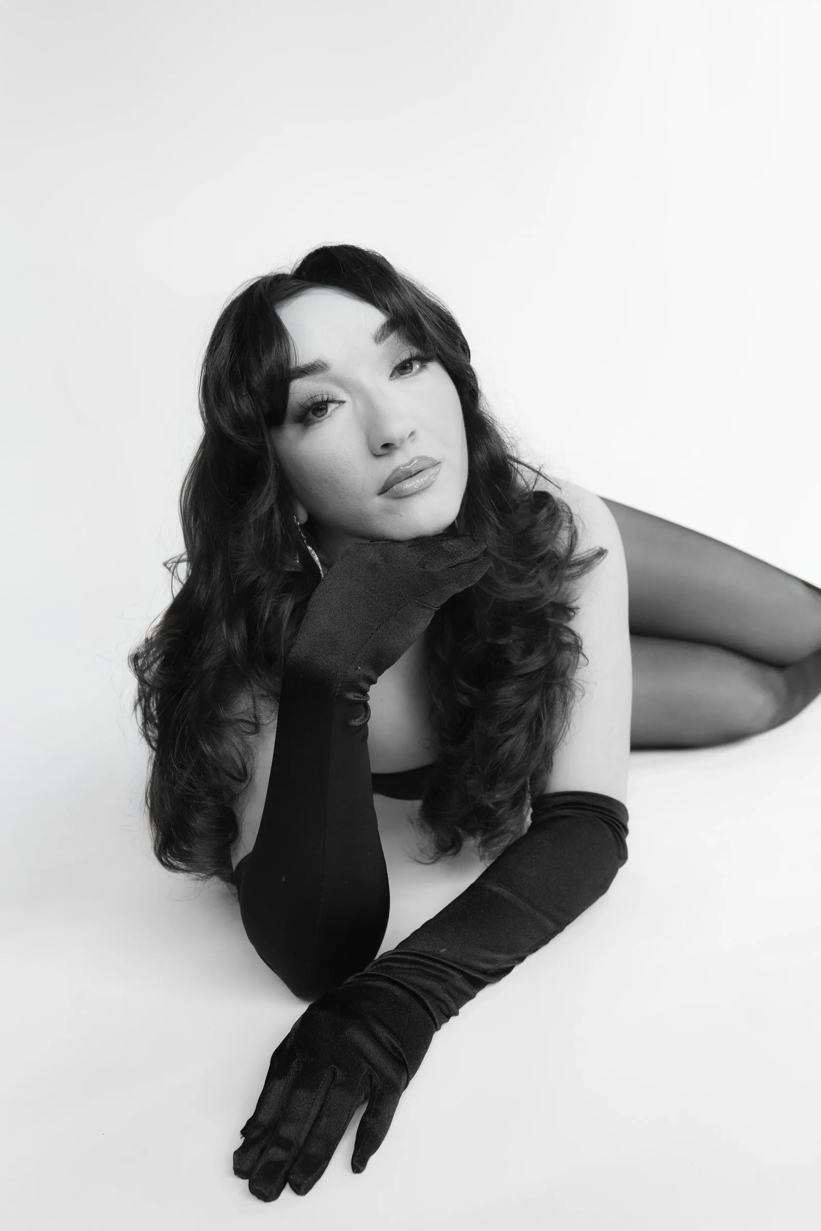 Black and white studio portrait of a woman with long curls in black opera gloves, seated on the floor