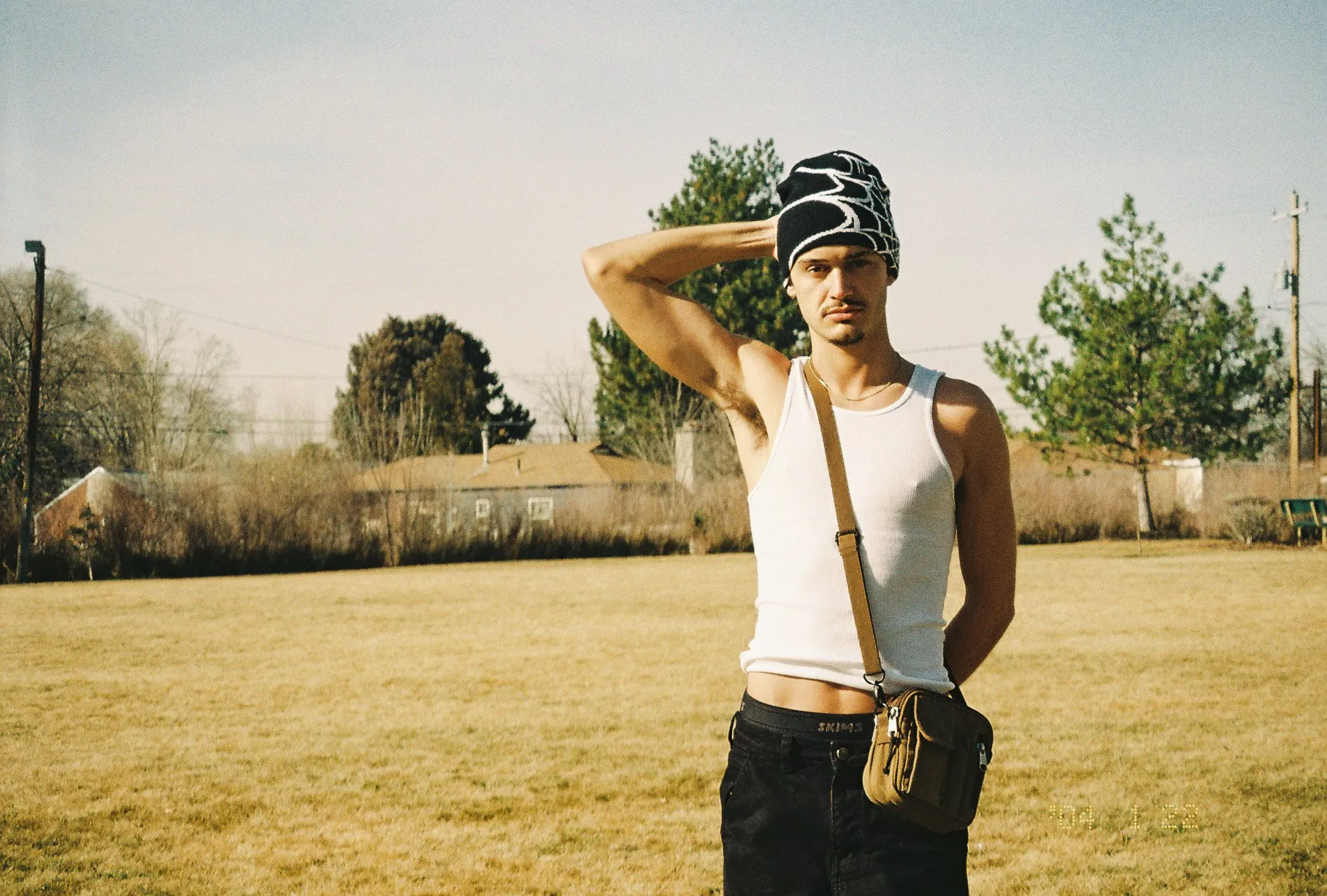 A man in a white tank top and patterned beanie stands in an open field, shot on film