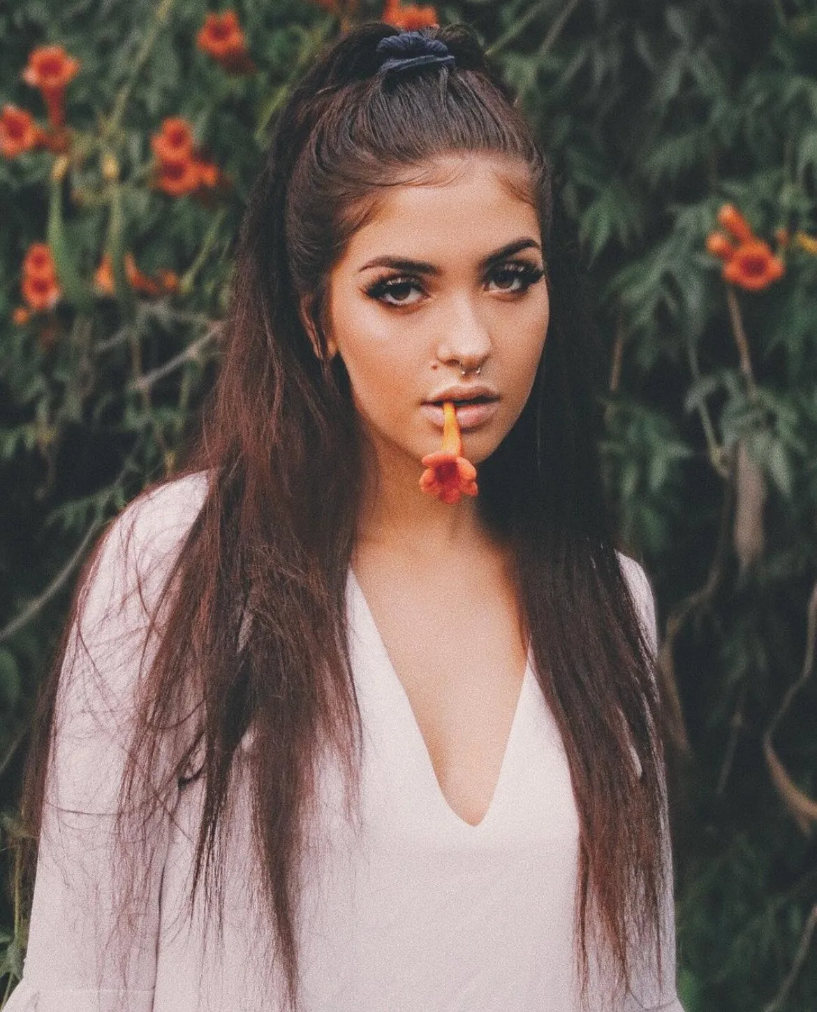 A woman with long dark hair holds an orange flower stem in her teeth in front of a flowering hedge