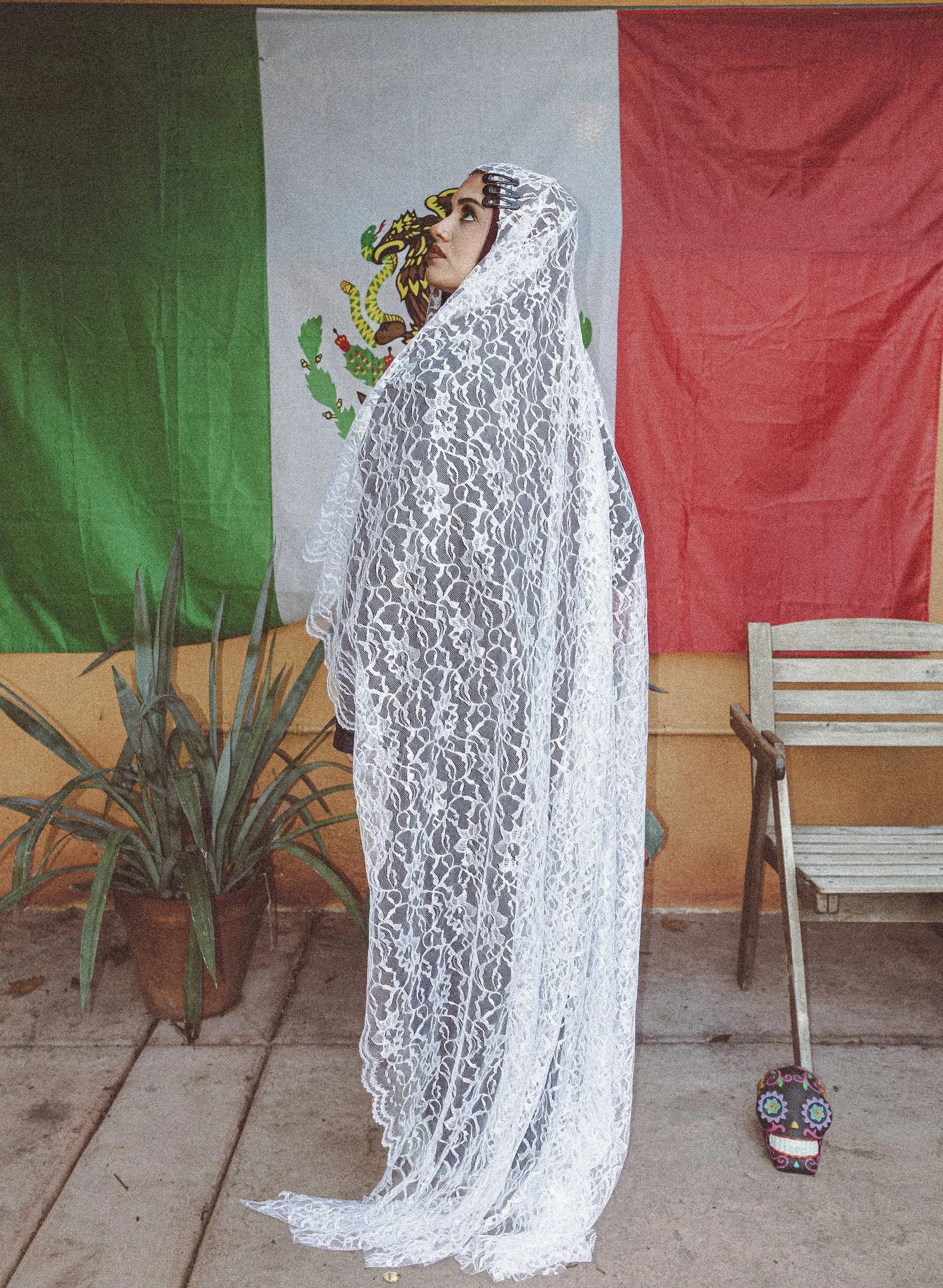 A figure draped head to toe in white lace stands before a Mexican flag on a sunlit patio