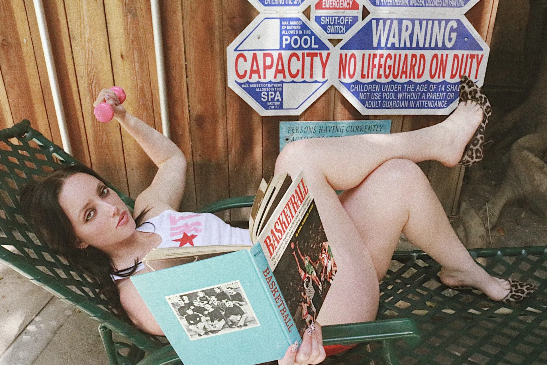 A woman reads a magazine on a green pool lounger beneath a fence covered in pool warning signs