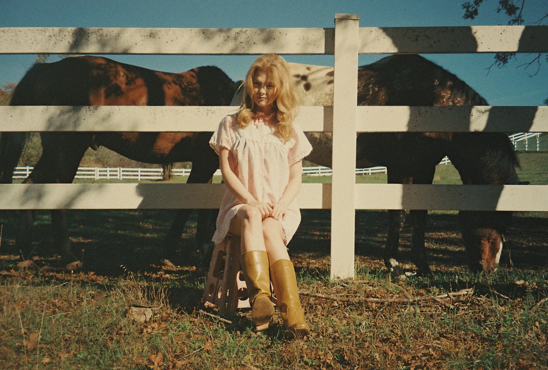 A woman in a pink slip dress and yellow boots sits by a white ranch fence as horses graze behind her