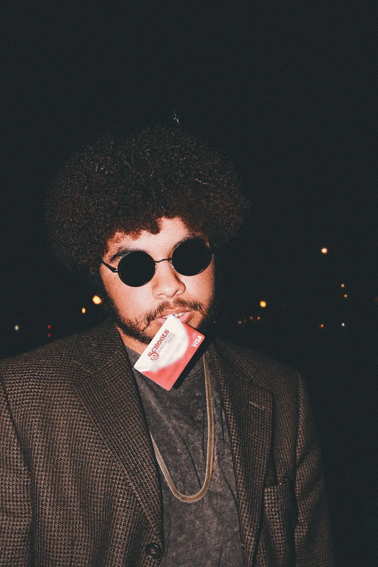 A man with an afro, round sunglasses and a tweed jacket holds a card in his teeth at night