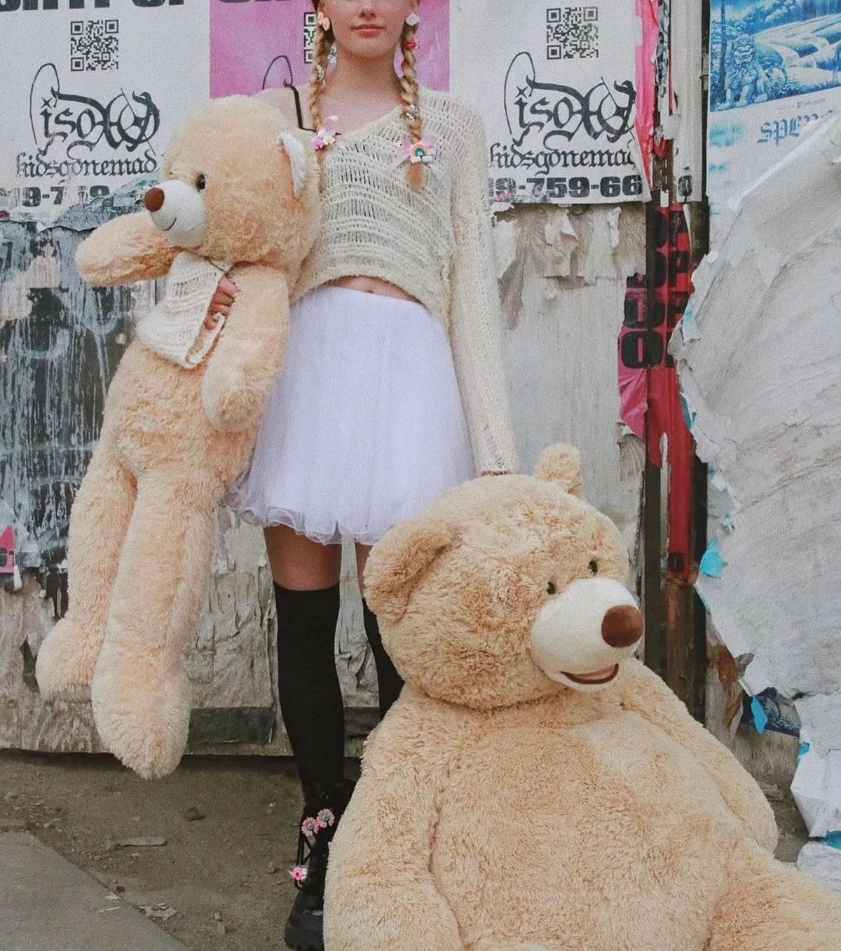 A model in a white tutu and knee socks stands with two giant teddy bears against a wall of street posters