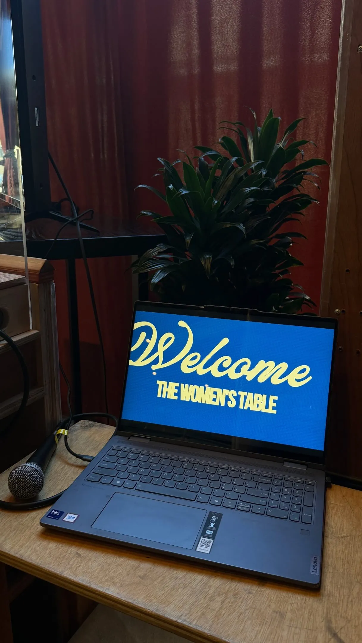 A laptop on a wooden table shows a welcome screen for The Women’s Table, the first Community Ties evening